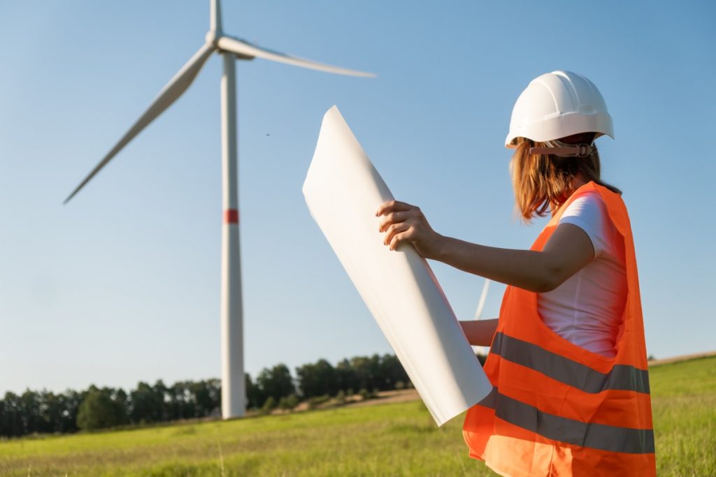 An engineer of wind turbines holding a paper project and looking at windmill in the field. Green eco