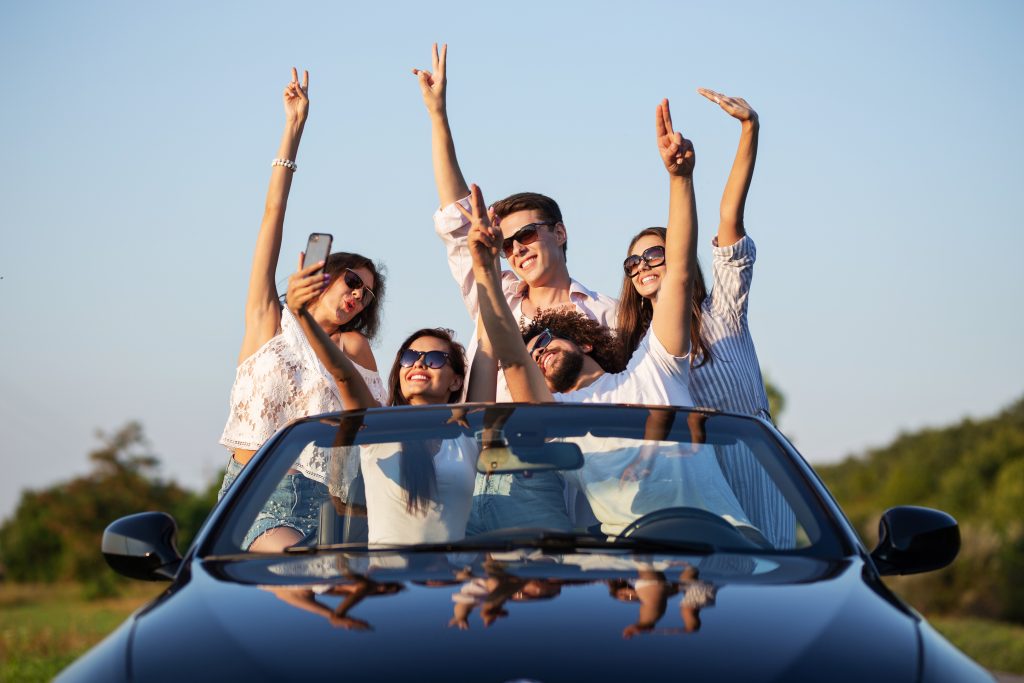 Young lucky girls and boys in sunglasses are sitting in a black cabriolet on the road holding their